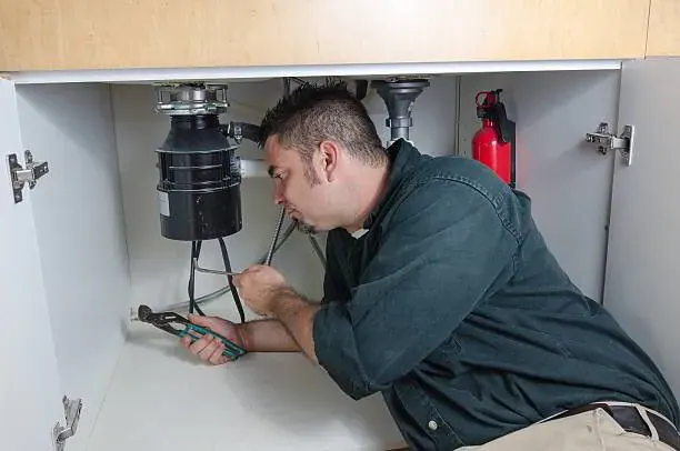 Plumber providing garbage disposal repair services beneath a kitchen sink